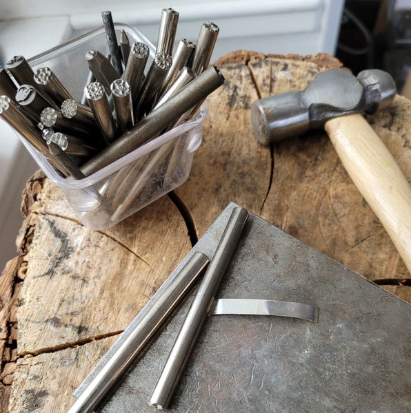 Metal stamps and hammer next to silver ring band on forging block.
