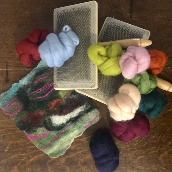 A pile of merino wool tops, hand held carders on top of a small wet felted abstract landscape