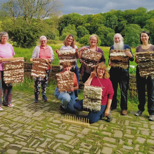 8 people holding up their woven mats, which include a range of creams, browns and greys. 