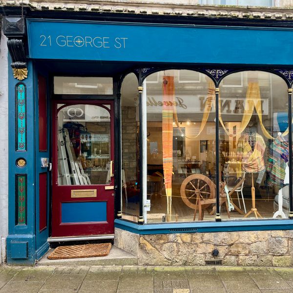 Colourful shop exterior with large sectional windows with curved tops. Painted in a dark turquoise with burgundy at bottom of the door and gold and turquoise embellishments painted onto upright section next to door. Inside the window you can see a spinning wheel and orange and yellow hanging strip of fabric