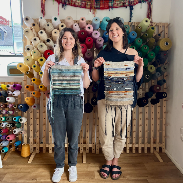 Two smiling women stand in front of a wall stand of colourful weaving cones. There is a window to the left. They both hold pieces of woven fabric infront of their chests in blues and natural tones. 